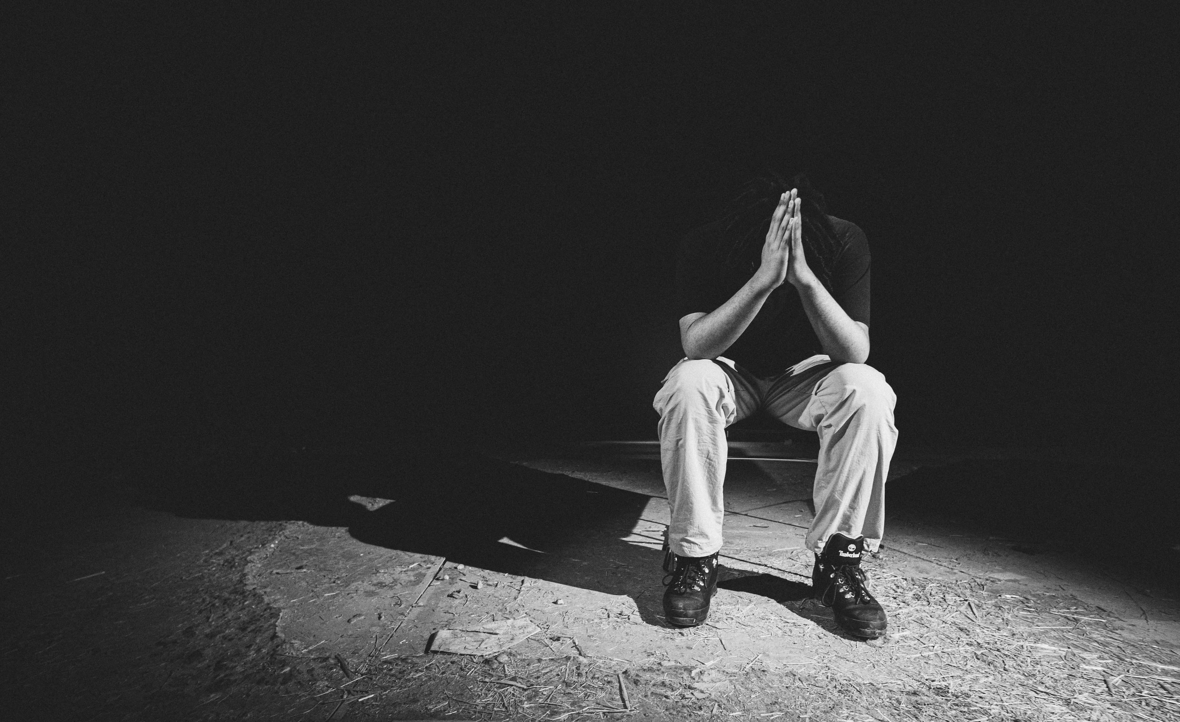 Person sitting alone in dim light, head bowed, hands clasped in quiet sorrow